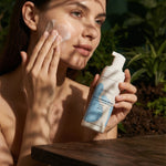 Woman applying skincare product to her face with a bottle of the Face Cleansing Mousse with Honeysuckle & White Willow product in her hand, outdoors.