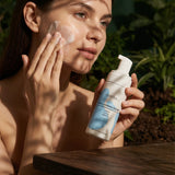 Woman applying skincare product to her face with a bottle of the Face Cleansing Mousse with Honeysuckle & White Willow product in her hand, outdoors.