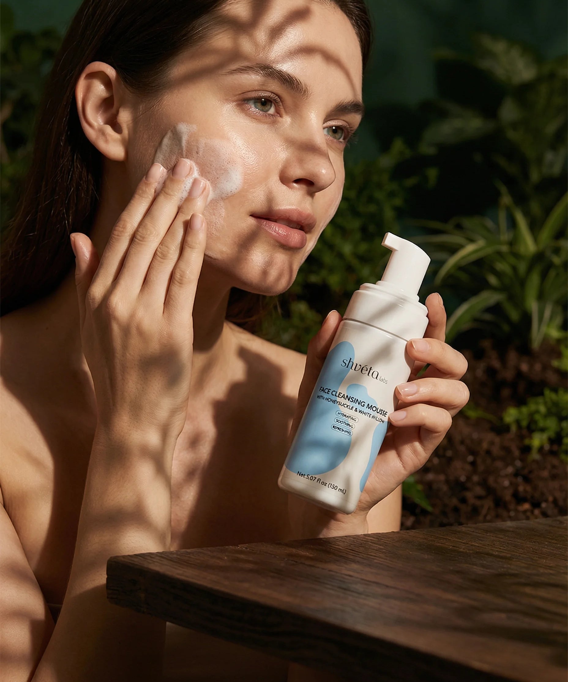 Woman applying skincare product to her face with a bottle of the Face Cleansing Mousse with Honeysuckle & White Willow product in her hand, outdoors.