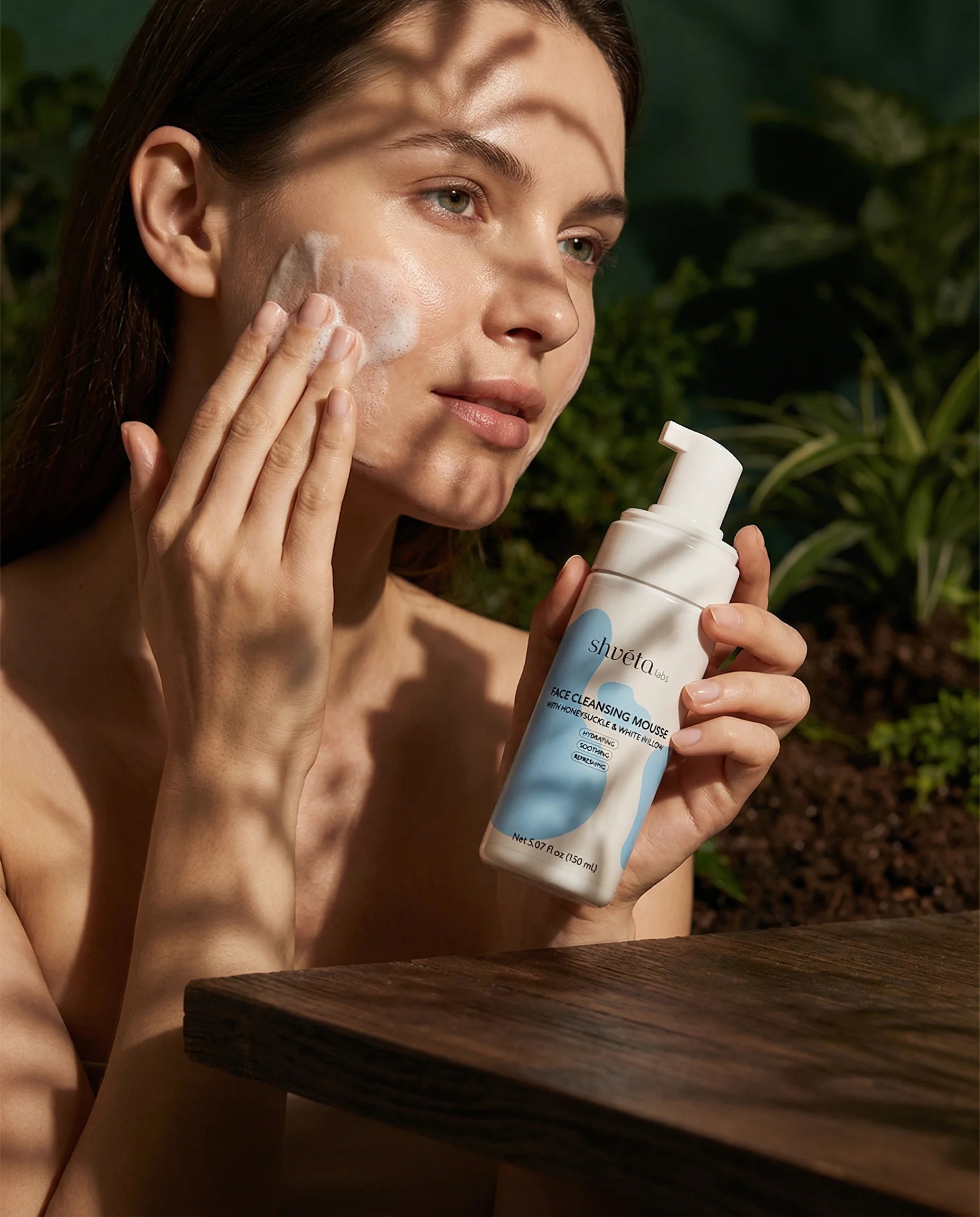 Woman applying skincare product to her face with a bottle of the Face Cleansing Mousse with Honeysuckle & White Willow product in her hand, outdoors.